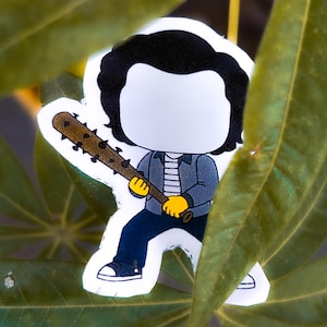 May include: A sticker featuring a cartoon character with black hair, a blue jacket, and blue pants, holding a brown baseball bat. The character has yellow gloves and black shoes. The sticker is set against a background of green leaves.
