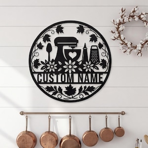May include: A circular black metal wall sign featuring a cutout design of a stand mixer, a spoon, a bottle, and a grater, surrounded by maple leaves and flowers. The text CUSTOM NAME is centered horizontally within the circular frame.