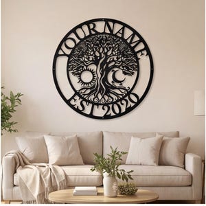 May include: A circular black metal wall art piece featuring a detailed tree of life design. The text YOUR NAME is arched across the top, while EST 2020 is arched along the bottom. A sun and a crescent moon are integrated into the tree branches.