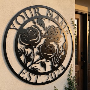 Puede incluir: Arte de pared de metal negro con un diseño circular. Las palabras "YOUR NAME" y "EST 2024" rodean un arreglo floral de rosas. La obra de arte está montada en una pared de color claro.