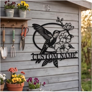 May include: A black metal wall sign featuring a hummingbird in flight next to three flowers, enclosed within a circular frame. Below the circular design, a horizontal bar displays the text CUSTOM NAME in bold, capital, sans-serif letters.