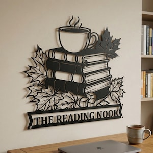 May include: Black metal wall art featuring a stack of books topped with a coffee cup, surrounded by maple leaves. The words "THE READING NOOK" are displayed on a banner below the books. A cozy, literary-themed decor piece.