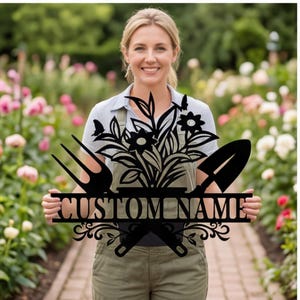 Puede incluir: Letrero de jardín de metal negro con el texto "CUSTOM NAME", con un diseño floral, mariposas, una horca de jardín y una paleta. El letrero lo sostiene una persona en un jardín con flores.