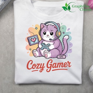 May include: White t-shirt featuring a cartoon cat wearing headphones and holding a game controller. The cat is purple with a pixelated heart on a tablet. The text "Cozy Gamer" is in red script.