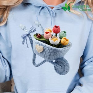 May include: Light blue hoodie featuring a crocheted wheelbarrow filled with colorful tulips. The wheelbarrow has a heart-shaped patch and a ladybug. The hoodie has the text "GraphiTee Charm" in the upper right corner.