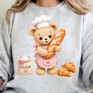 May include: A light gray sweatshirt featuring a graphic of a crocheted teddy bear wearing a white chef hat and a pink gingham apron. The bear holds a baguette, with a sack labeled FLOUR and two croissants positioned nearby on the fabric.