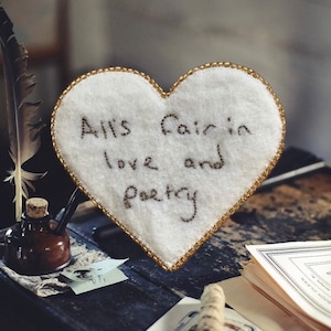 May include: A white heart-shaped pillow with gold trim and the embroidered text "All's fair in love and poetry".