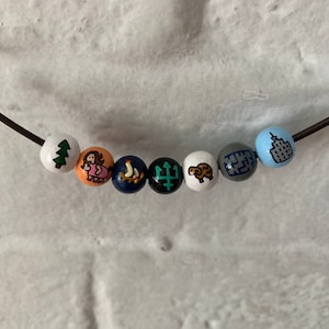 Puede incluir: Un collar de cuentas blancas con varios s&iacute;mbolos pintados, incluyendo un &aacute;rbol, una persona, un barco, un tridente, un carnero, un edificio y un horizonte de la ciudad.