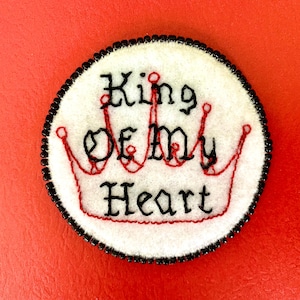 May include: A white felt circle with a black beaded border, featuring a red embroidered crown and the text "King of My Heart".
