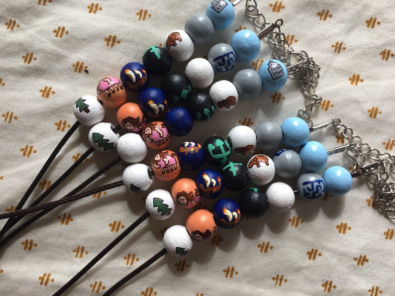 Puede incluir: Un conjunto de cuentas de madera de colores con varios dise&ntilde;os, incluyendo &aacute;rboles, animales y formas geom&eacute;tricas. Las cuentas est&aacute;n enhebradas en cordones negros y algunas tienen cierres plateados.