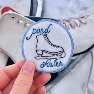 May include: A blue and white embroidered patch with the text "pond skater" and a black and white illustration of an ice skate.