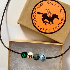 May include: A brown cord necklace with four painted wooden beads. The beads feature a trident, a ram, a maze, and a building. The necklace is displayed with a small cardboard box with the words "BLOOD CAMP HALF-BLOOD" and a winged horse logo.