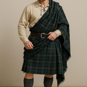 Könnte beinhalten: Traditionelles schottisches Kilt-Outfit. Das Ensemble umfasst einen grün-rot karierten Kilt, einen passenden Schal, ein beiges Hemd, einen braunen Gürtel, graue Socken und schwarze Schuhe. Das Outfit wird von einer Person mit Bart getragen.