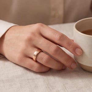 May include: A smooth, polished rose gold band ring with a simple, rounded profile. The metal has a warm, metallic pinkish hue and a reflective surface, worn on a finger next to a ceramic mug containing a dark liquid on a light-colored surface.