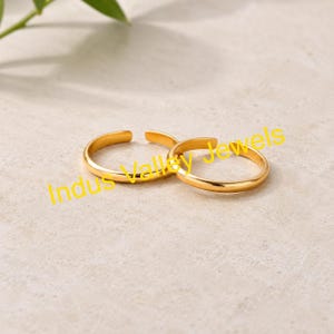 May include: Two gold-coloured adjustable rings are displayed on a light-coloured surface. The rings are open-ended, allowing for size adjustment. The rings are simple, with a smooth, polished finish. The rings are likely jewellery.