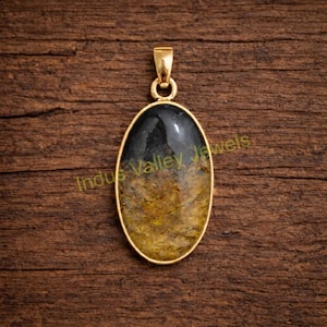 May include: Oval pendant with a gold-colored frame and a polished stone. The stone has a dark gray top and a yellow-brown bottom. The text "Indus Valley Jewels" is printed on the stone.