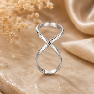 Könnte beinhalten: Ein silberfarbener Ring mit einem unendlichkeitsförmigen Design und einer gehämmerten Textur. Das Band kreuzt sich in der Mitte zu zwei Schlaufen und bildet eine durchgehende Acht-Silhouette mit einer polierten, metallisch glänzenden Oberfläche.