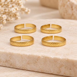 Könnte beinhalten: Vier goldfarbene Metallringe, die in einer quadratischen Formation angeordnet sind. Jeder Ring verfügt über ein offenes Banddesign mit einem strukturierten, schraffierten Muster auf der Außenseite. Die Ringe haben eine polierte Oberfläche.