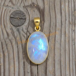 May include: Oval moonstone pendant with a gold-colored setting and bail. The moonstone has a blue and white iridescent glow. The pendant is displayed on a wooden surface.