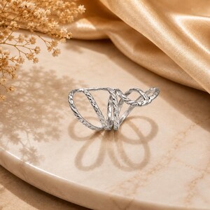 May include: A silver-toned multi-band ring featuring a textured, patterned surface across its interconnected loops. The design consists of several curved, overlapping wire-like segments that create an open, airy structure with a polished metallic finish.