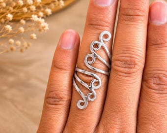 Puede incluir: Un anillo de metal plateado con un diseño alargado que cubre una parte importante del dedo. El anillo está compuesto por múltiples elementos de alambre curvos y en espiral que crean una serie de bucles abiertos interconectados.