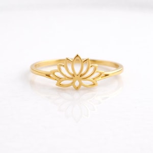 May include: A gold-colored ring featuring a lotus flower design. The ring's band is thin, and the lotus flower is the focal point, with its petals delicately outlined. The ring is set against a white background, highlighting its details.