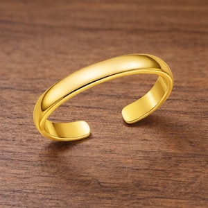 May include: A gold-coloured toe ring with a smooth, curved design. The open-ended ring has a polished finish, reflecting light. The jewellery is set against a warm, wooden surface.