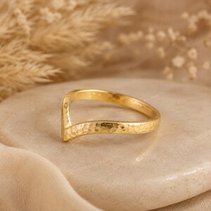 Op de afbeelding: Een goudkleurige ring met een gehamerde textuur en een V-vormig ontwerp. De ring ligt op een neutrale ondergrond, met een vage achtergrond. Het ontwerp van de ring is eenvoudig maar elegant, geschikt voor dagelijks gebruik.