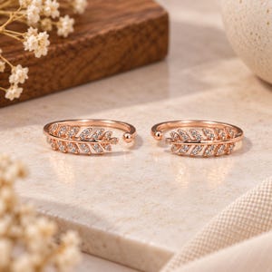 Könnte beinhalten: Zwei offene Ringe in Roségold-Optik mit einem filigranen Blatt-Design. Jeder Ring ist mit kleinen, klaren, funkelnden Steinen besetzt, die entlang der blattartigen Struktur angeordnet sind und ein zweigartiges Aussehen erzeugen.
