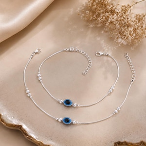May include: Two silver anklets with a delicate chain design. Each anklet features a blue and black evil eye charm and small silver beads. The anklets are displayed on a beige surface, with dried flowers in the background.