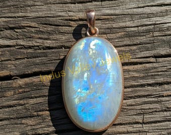 May include: Oval-shaped pendant with a large, iridescent moonstone. The stone has a pearly white base with blue and iridescent flashes. The pendant is set in a rose gold-colored metal with a simple bail. The background is a weathered wooden surface.