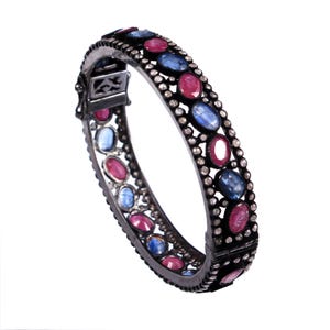 Può includere: Un bracciale rigido incernierato con una montatura in metallo scuro incastonata con pietre preziose ovali alternate blu e rosa. I bordi del bracciale sono delineati da piccole pietre rotonde e chiare, creando un bordo dettagliato e strutturato.
