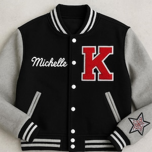Op de afbeelding: Zwarte varsity jas met grijze mouwen. Voorzien van de naam "Michelle" in wit script en een grote rode "K" met een witte omtrek. De jas heeft wit-zwarte gestreepte manchetten en een sterpatch op de mouw.