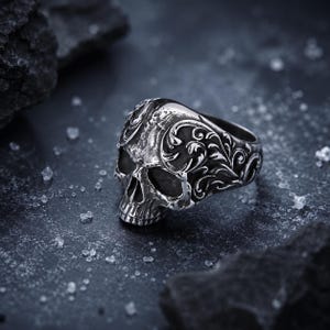 May include: A silver-toned metal ring shaped like a human skull. The surface of the ring features intricate, raised scrollwork and filigree patterns carved into the metal, with darkened recessed areas to highlight the detailed texture of the skull.