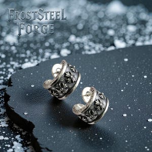 May include: Two silver hoop earrings with a floral pattern. The earrings are displayed on a dark surface, with the text "FrostSteel Forge" visible. The earrings have a curved design and are decorated with small, raised floral motifs.