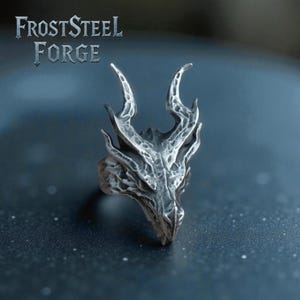 Könnte beinhalten: Ein silberner Ring in Form eines Drachenkopfes. Der Ring weist komplizierte Details auf dem Gesicht, den Hörnern und den Schuppen des Drachen auf. Der Text "FROSTSTEEL FORGE" wird über dem Ring angezeigt. Dunkler Hintergrund.