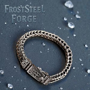 May include: A silver bracelet with a woven chain design and an ornate clasp. The bracelet is displayed on a dark blue surface with the text "FrostSteel Forge". The clasp features intricate patterns. The bracelet is made of silver.