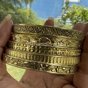 May include: A stack of gold-colored metal bangles with intricate floral and geometric patterns. The bangles have a textured surface and are stacked together. The text "PRhandicraft" is visible on the bangles.