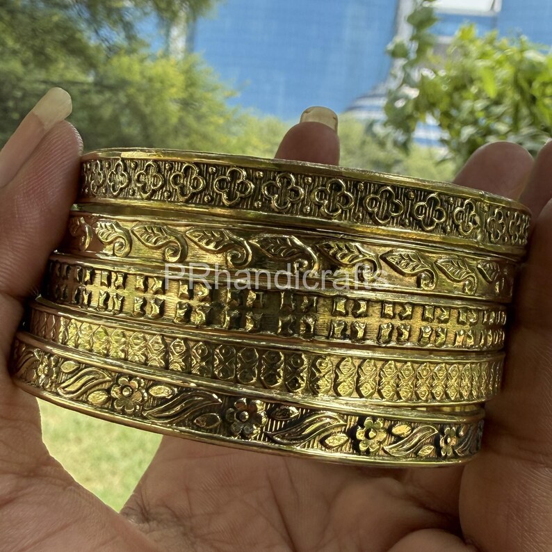 May include: A stack of gold-colored metal bangles with intricate floral and geometric patterns. The bangles have a textured surface and are stacked together. The text "PRhandicraft" is visible on the bangles.