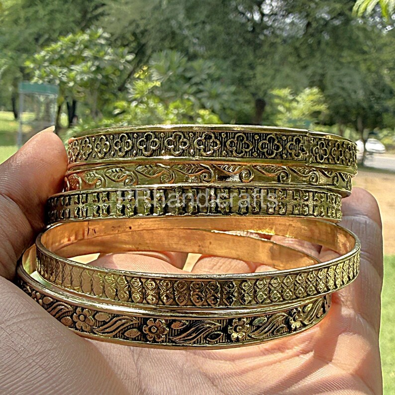 May include: A stack of gold-colored bangles with intricate floral and geometric patterns. The bangles are stacked in a hand, showcasing the detailed designs and textures. The image is taken outdoors with a blurred green background.