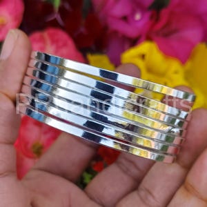 May include: A stack of silver-colored bangles. The bangles are thin, flat, and shiny, reflecting light. The background features a blurred mix of colorful flowers, including pink, red, and yellow blooms. The bangles are held in a hand.