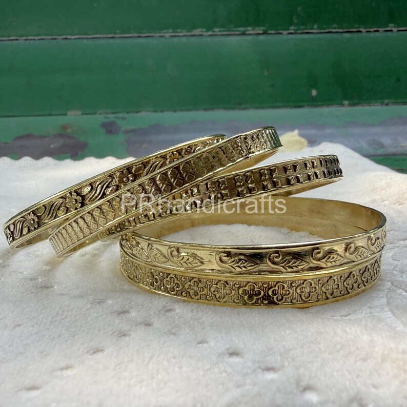 May include: Four gold-colored bangles with intricate floral and geometric patterns. The bangles are stacked on a white surface, showcasing their detailed designs and polished finish. The bangles are a classic jewelry item.