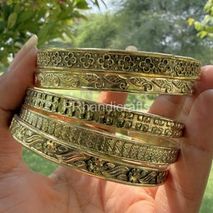 May include: A stack of gold-colored bangles with intricate floral and geometric patterns. The bangles are held in a hand, showcasing their detailed design and polished finish. The text "PRhandicrafts" is visible on the bangles.