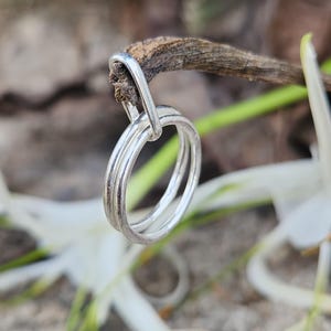 Puede incluir: Un anillo plateado con tres bandas conectadas cuelga de una pequeña ramita marrón. El anillo está enfocado, con un fondo borroso de flores blancas y vegetación. El anillo tiene un diseño simple y elegante.