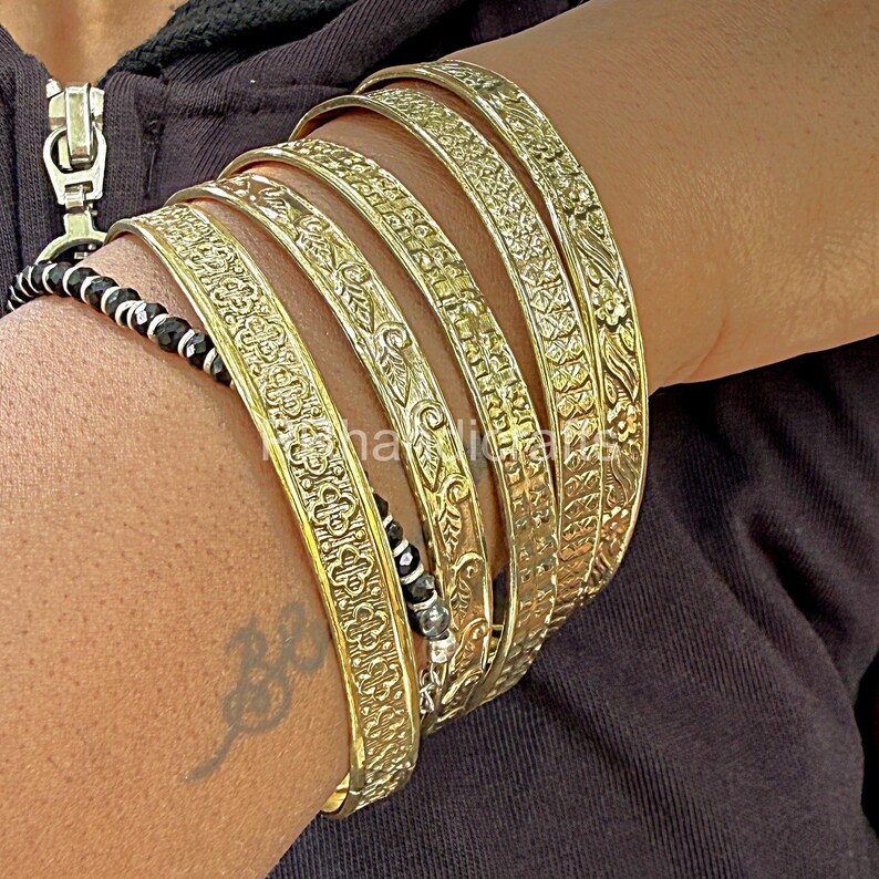 May include: A collection of gold-colored bangles with intricate floral and geometric patterns. The bangles are stacked on a wrist, showcasing their detailed designs. A black and white beaded bracelet is also visible.