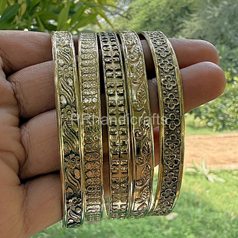 May include: A set of six gold-colored bangles with intricate floral and geometric patterns. The bangles are held in a hand, showcasing the detailed designs. The image includes the text "RKhandicrafts".