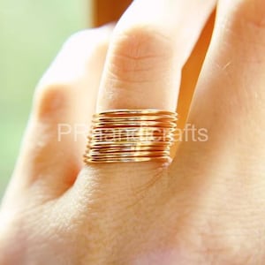 Op de afbeelding: Een stapel dunne, goudkleurige ringen die op een vinger worden gedragen. De ringen zijn dicht op elkaar gepakt, waardoor een textuur ontstaat. De achtergrond is wazig, met zacht, natuurlijk licht.