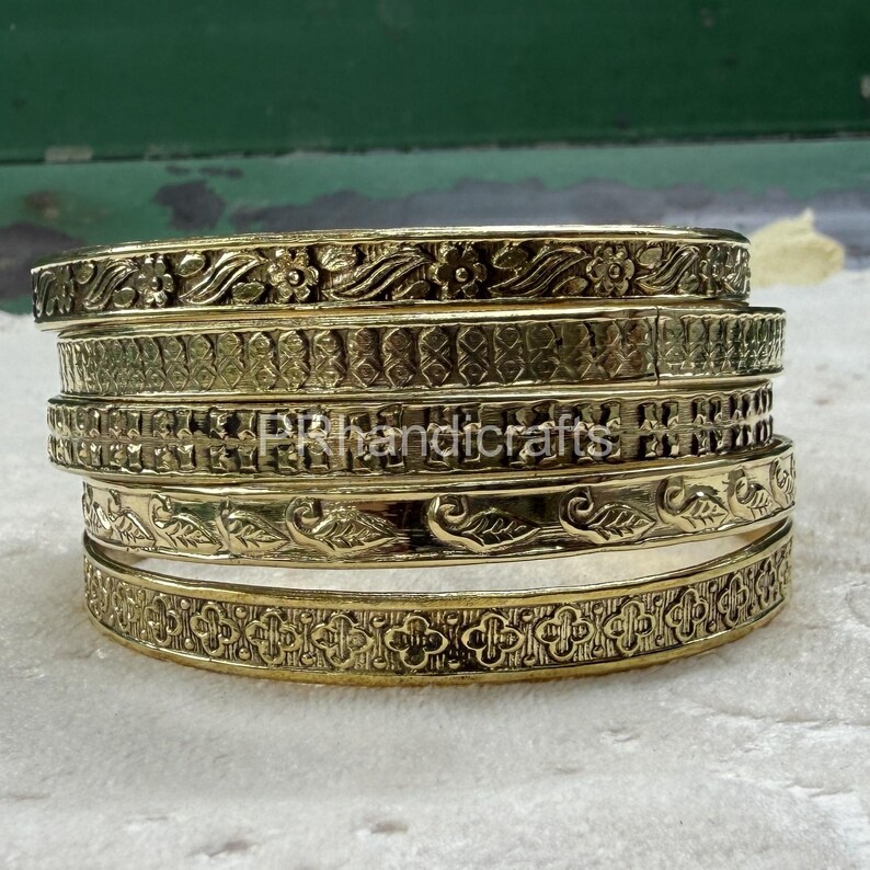 May include: A stack of six gold-colored bangles with intricate floral and geometric patterns. The bangles are stacked on top of each other, showcasing the detailed designs. The text "PR handicrafts" is visible on one of the bangles.