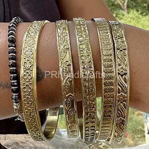 May include: A collection of gold-colored bangles with intricate floral and geometric patterns. The bangles are worn on a wrist alongside a black beaded bracelet. The bangles have a width of approximately 0.5 inches.