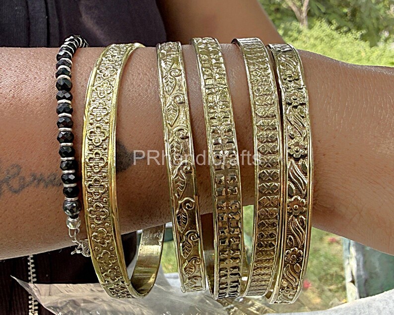 May include: A collection of gold-colored bangles with intricate floral and geometric patterns. The bangles are worn on a wrist alongside a black beaded bracelet. The bangles have a width of approximately 0.5 inches.